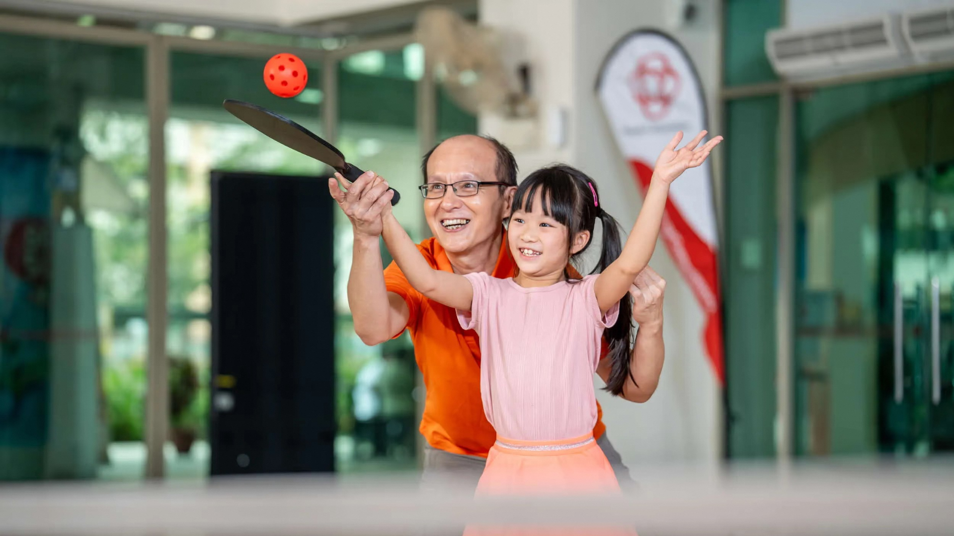 A person in an orange shirt helps a child in a pink shirt and orange skirt hit a ball with a paddle indoors.